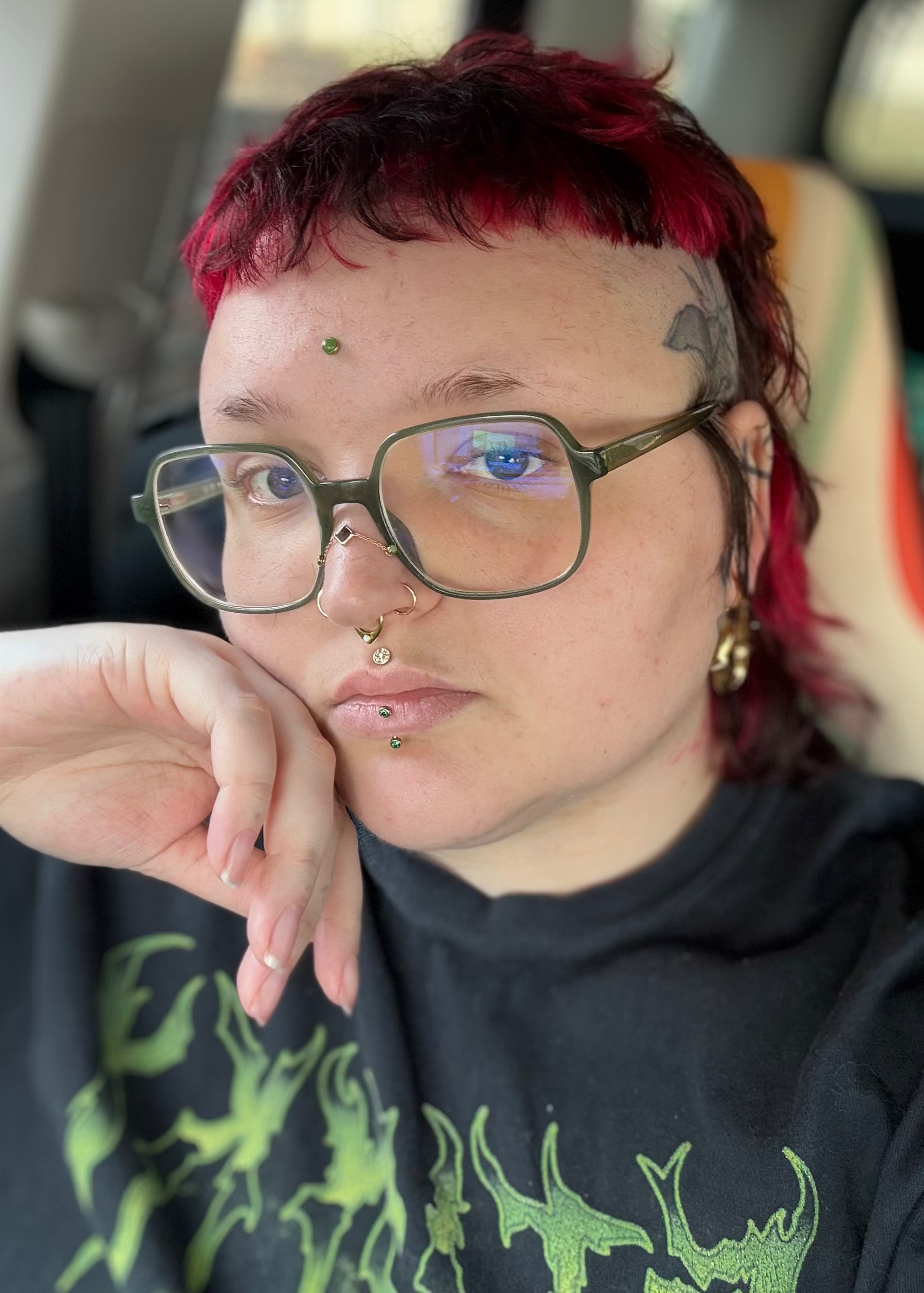 A androgynous person with a short red hair, several facial piercings, and green glasses sitting inside a vehicle.